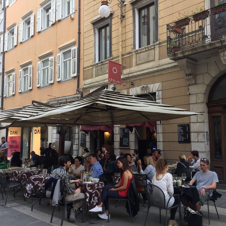 I tavoli all'aperto del New Circus in via San Lazzaro a Trieste, sotto gli ombrelloni a righe, con l'insegna Circus e le lavagne del menu